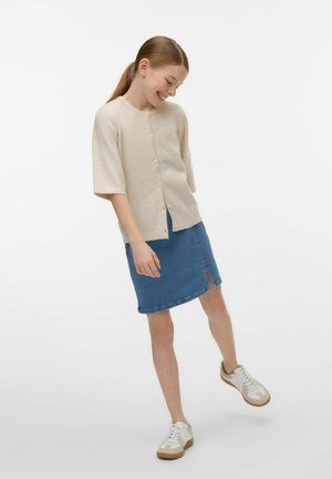 Jonge vrouw die glimlacht, gekleed in een beige gebreid vest met knopen, een spijkerrok met voorste split en witte sneakers, staande tegen een effen achtergrond.