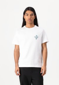T-shirt en coton blanc avec un col rond, des manches courtes et un petit graphique de palmier teal et jaune dans un design triangulaire sur la poitrine.