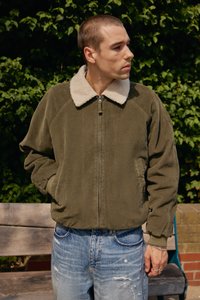 Chaqueta de corduroy verde oliva con un cuello de piel crema, textura acanalada, cierre de cremallera y bolsillos laterales, combinada con jeans azules desgastados.