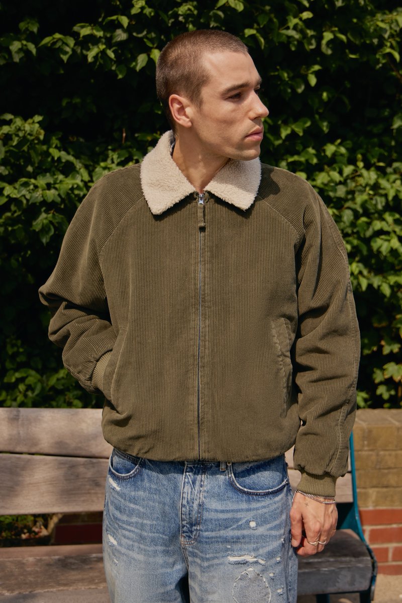 Chaqueta de corduroy verde oliva con un cuello de piel crema, textura acanalada, cierre de cremallera y bolsillos laterales, combinada con jeans azules desgastados.
