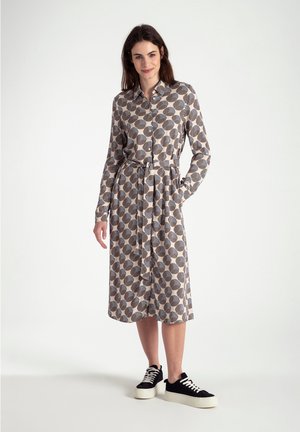 Knopfleistenkleid in hellem Beige mit einem kreisförmigen Muster in Dunkelbraun und Streifen, in der Taille mit einem Bindegürtel gerafft, kombiniert mit schwarzen Plateausneakern.