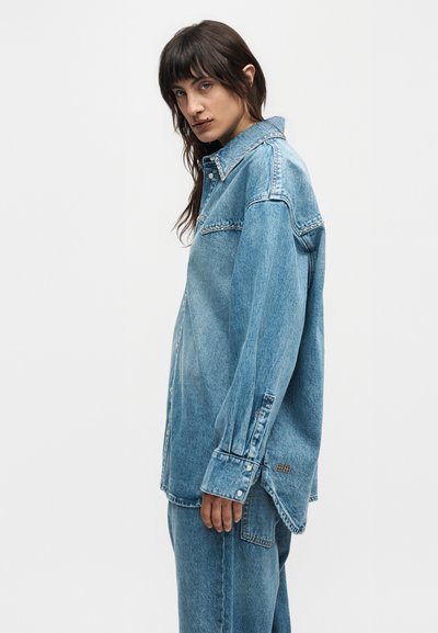 Γυναίκα με μακριά σκούρα μαλλιά, φορώντας υπερμεγέθη ανοιχτό μπλε τζιν πουκάμισο και τζιν παντελόνι που ταιριάζει, στέκεται πλαγίως μπροστά σε ένα απλό φόντο.
