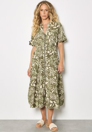 Kobieta ubrana w zieloną, midi sukienkę na zamek z przodu z białymi wzorami liści i krótkimi rękawami, w połączeniu z beżowymi sandałami wsuwanymi.