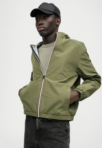 Veste coupe-vent vert olive avec fermeture éclair sur le devant, poignets élastiques et deux poches latérales. Elle possède une doublure intérieure contrastante et une fermeture éclair rayée.