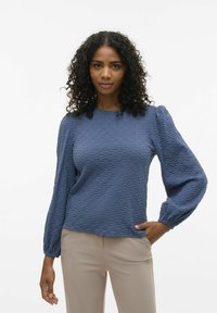 Maglia lunga blu texturizzata con scollatura a girocollo e spalle a palloncino, abbinata a pantaloni beige. Il tessuto presenta un motivo ondulato e una leggera lucentezza.
