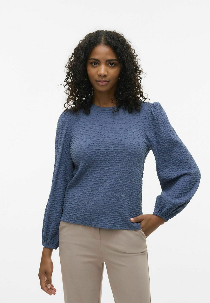 Maglia lunga blu texturizzata con scollatura a girocollo e spalle a palloncino, abbinata a pantaloni beige. Il tessuto presenta un motivo ondulato e una leggera lucentezza.