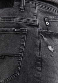 Jeans gris oscuro con un detalle desgastado en el bolsillo delantero y una etiqueta de logo sutil. Hechos de denim con una textura suave.