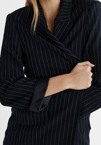 Marineblauwe blazer met krijtstreepdessin, dubbele knoopsluiting, zwarte satijnen manchetten en een gestructureerde stof. Voorzien van een reverskraag en zijzakken.