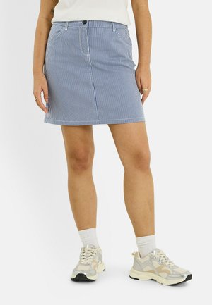 Mujer con falda de rayas verticales azul y blanca, calcetines blancos, zapatillas de colores claros y una blusa blanca sobre un fondo liso.