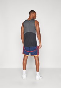 Sporty tank top i grå og sort på model, parret med blå shorts med røde accenter og gule kanter. Modellen bærer hvide sportssko.