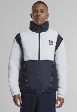 Mann trägt eine weiße und marineblaue Steppjacke mit hohem Kragen und Reißverschluss sowie Kani-Logo auf der Brust, stehend vor einem schlichten grauen Hintergrund.