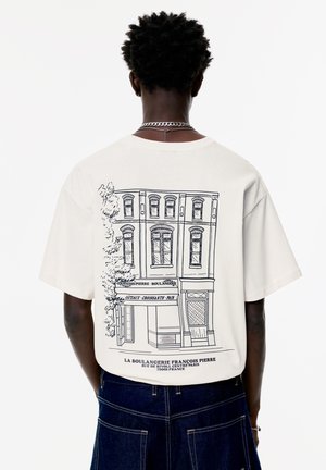 Cremefarvet bomulds-t-shirt med en sort stregtegning af et bageri med træer, der fremhæver designet på ryggen, løstsiddende pasform, korte ærmer.