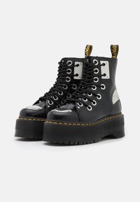 Botines de plataforma negros hechos de material brillante con acentos de metal plateado, punta redonda, suela gruesa y costura amarilla a lo largo del borde.