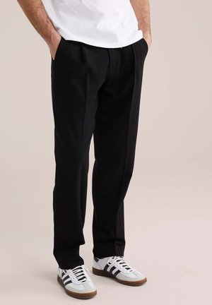 Mees, kes kannab musti pükse, käed taskutes ja valged tossud mustade triipudega beežil taustal.