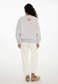 Šedá mikina s voľným strihom, rebrovanými manžetami a spodným okrajom. Na zadnej strane sa nachádza malá červená nášivka s logom. Nosená s svetlými nohavicami.