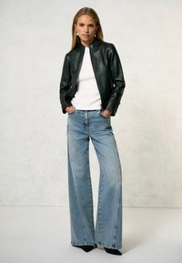 Veste en cuir noire au design court, t-shirt blanc ajusté en dessous, jean en denim bleu clair à jambes larges, et chaussures noires à bout pointu.