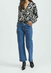 Schwarze Bluse mit lila und cremefarbenen abstrakten Mustern, kombiniert mit blauen Hochbundjeans mit geradem Bein. Getragen mit spitzen schwarzen Knöchelstiefeln.