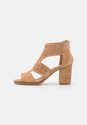 Sandale à talon ouverte en daim couleur camel avec motifs géométriques découpés, talon bloc et fermeture éclair arrière sur fond blanc.