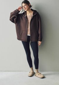 Overdimensioneret brun zip-up hættetrøje med hætte, parret med sorte leggings og beige træklip. Gulvlængde med lommer og ribbet detaljering på ærmerne.