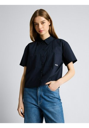 Młoda kobieta w czarnej, krótkorękawowej koszuli na guziki i niebieskich dżinsach, stojąca z jedną ręką w kieszeni na jednolitym tle.