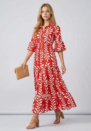 Mujer vistiendo un vestido maxi floral rojo y blanco con mangas con volantes, sosteniendo un bolso de mano beige y llevando tacones gruesos con tiras color cuero.