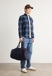 Camisa de flanela xadrez em azul-marinho, t-shirt branca, jeans azul-claro, segurando uma mala de viagem azul-marinho, usando um boné castanho e ténis brancos.