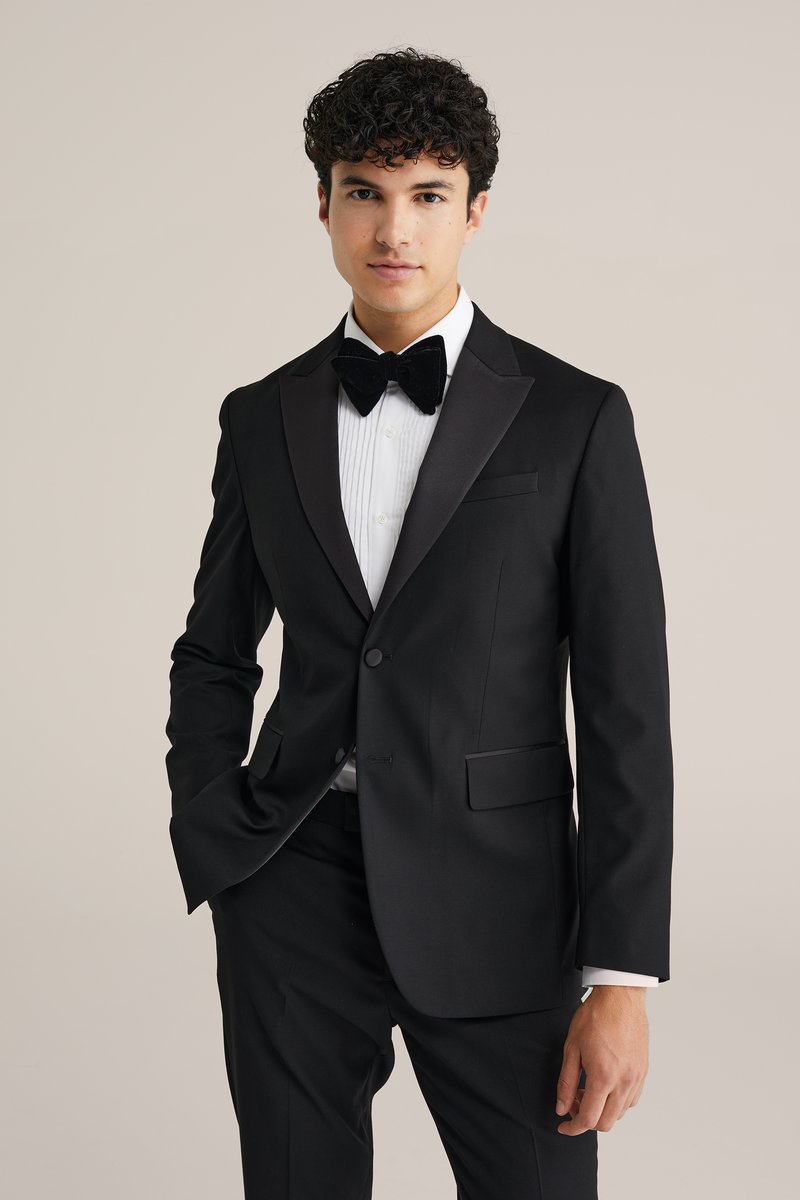 Črn smoking s satenskimi reverji in belim nabranim srajcem. Vključuje črno metuljčkasto kravato. Gladek material z krojem po meri.