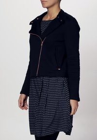 Veste noire cintrée avec fermeture éclair à l'avant, dotée d'un col et d'accents côtelés aux manches. Portée sur une robe à motifs noir et blanc.