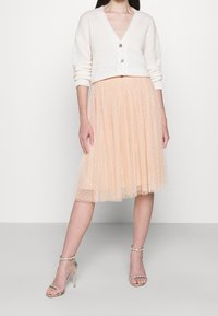 Cardigan beige con scollo a v e due bottoni argento, abbinato a una gonna in tulle pesca con pois e scarpe con tacco a strappo argento.