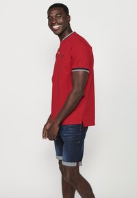 Tricou polo roșu, cu o țesătură texturată, având un guler îngust și accente cu dungi pe mâneci. Asortat cu blugi scurți.