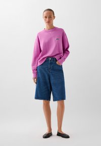 Hanorac roz supradimensionat cu logo brodat, asortat cu pantaloni scurți din denim albastri până la genunchi. Modelul poartă pantofi slip-on negri.