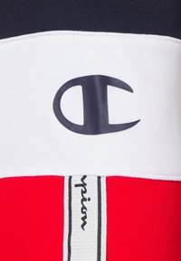Punainen ja valkoinen urheiluvaate, jossa on suuri musta logo ja pystysuora "Champion" -merkki tekstuurilla varustetussa taustakuosissa.