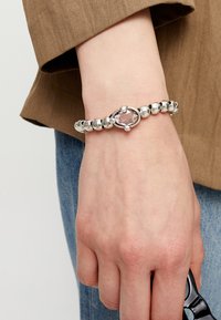 Pulsera de plata con cuentas esféricas y una piedra ovalada morada como pieza central, llevada en una mano sobre un fondo de manga marrón y mezclilla.