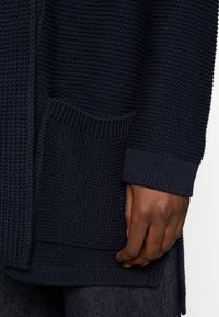 Cardigan in maglia blu marino con un motivo testurizzato, dotato di una tasca laterale e polsini a costine. Il tessuto è spesso e leggermente elasticizzato.
