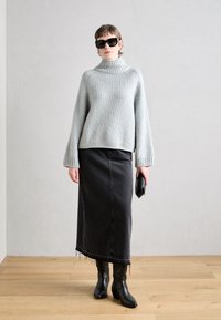 Světlé šedé pletené svetr s vysokým límcem, černá džínová sukně až po střed lýtek a černé kožené boty. Drží černou kabelku. Jednoduché pozadí.