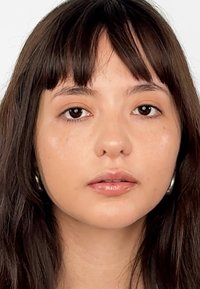 Ritratto di una persona con lunghi capelli scuri e pelle di un marrone chiaro, con trucco delicato e grandi orecchini a cerchio in argento, su uno sfondo semplice.