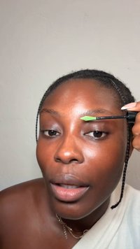 Frau mit geflochtenem Haar, die grünes Augenbrauengel auf ihre rechte Augenbraue aufträgt vor einem einfarbigen hellen Hintergrund.