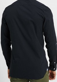 Mörkblå långärmad skjorta med en slät textur, med en avslappnad passform och knappdetaljer på ärmsluten, sedd bakifrån.