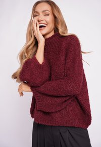 Burgundy knit sweater with a high collar, textured fabric, and oversized sleeves, paired with a pleated black skirt.