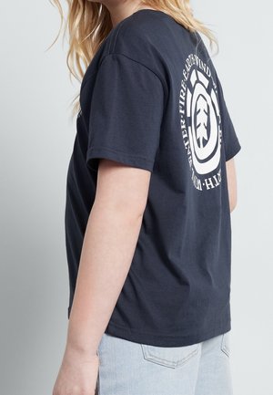 Personne portant un t-shirt bleu marine avec un logo circulaire blanc et un texte au dos, associé à un jean en denim bleu clair, debout devant un fond uni.