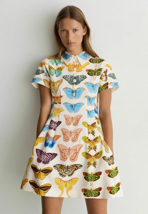 Vrouw draagt een witte jurk met korte mouwen, kleurrijke vlinderprints, een Peter Pan-kraag en knopen aan de voorkant, staande tegen een eenvoudige muur.
