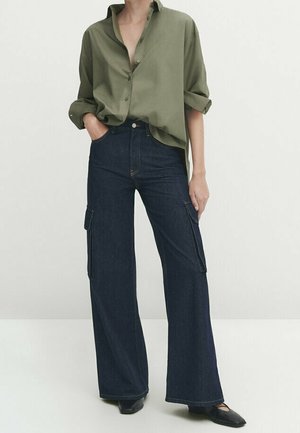 Person wearing olive green buttoned shirt with rolled sleeves and dark blue wide-leg cargo jeans with pockets and black shoes.