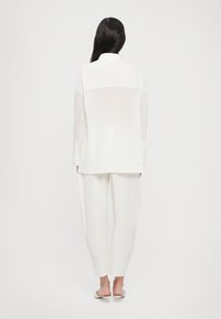 Hvit, lett bluse med høy-lav skjæring og lange ermer, kombinert med løst sittende hvite bukser. Glatt tekstur, minimalt design.