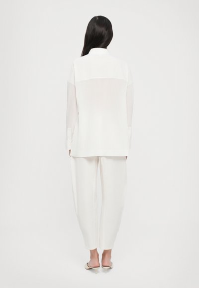 Blusa bianca leggera con orlo asimmetrico e maniche lunghe, abbinata a pantaloni bianchi a vestibilità ampia. Tessuto morbido, design minimalista.