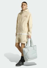Chaqueta deportiva beige y pantalones cortos con logotipo tonal; bolso tote gris con doble asa; zapatillas de deporte texturizadas; diseño minimalista.
