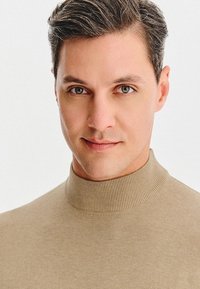 Mężczyzna z krótkimi, teksturowanymi szarymi włosami, noszący beżowy sweter z golfem. Tło jest jednolicie białe, co uwydatnia cechy postaci.