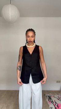 Άτομο με πλεξούδες, φοράει μαύρο αμάνικο γιλέκο, λευκό φαρδύ παντελόνι, στέκεται σε εσωτερικό χώρο με τατουάζ στο αριστερό χέρι και μεγάλο κολιέ.