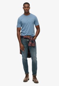 Camiseta azul de manga corta con logo, combinada con jeans de mezclilla, botas marrones y una camisa de cuadros anudada a la cintura.