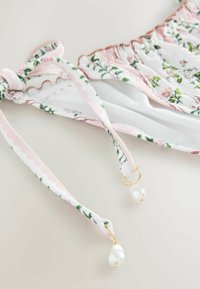Top de bikini floral cu bază albă și modele în roz și verde, margini cu volane și elemente aurii cu accente de perle pe bretele.