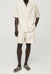 Hellbeiges kurzärmliges Hemd und passende Shorts aus strukturiertem Stoff. Die Schuhe sind braune Sandalen mit gekreuzten Riemen.